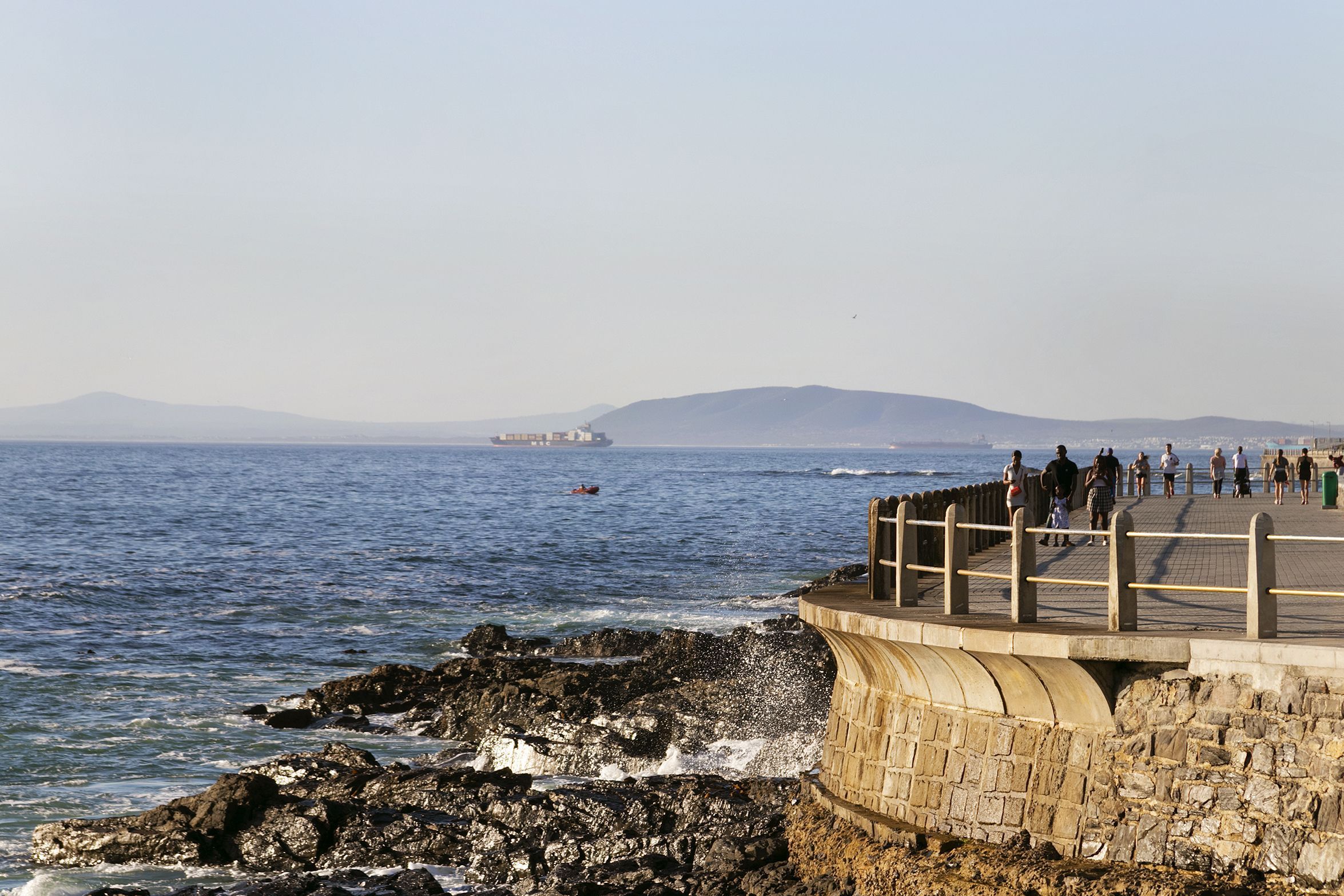 A coastal walkway with a stone barrier, a distant ship, and a mountainous backdrop.