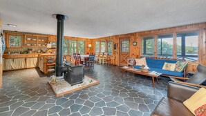 A cozy cabin interior with a stone floor, wooden walls, a central fireplace, a dining area, and large windows offering a view of the outdoors.