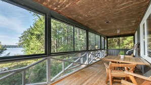 A covered deck with a wooden table and chairs, large windows offering a view of a forest and lake.