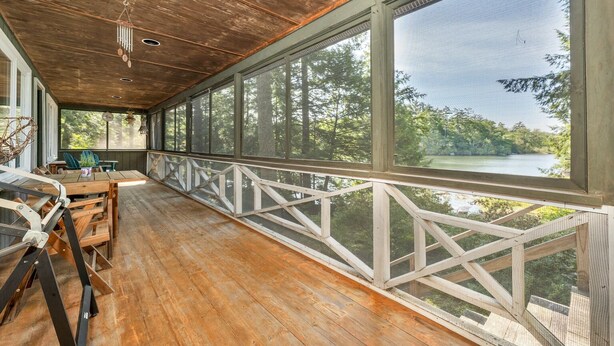 A covered porch with wooden flooring, large windows, and a view of a lake.