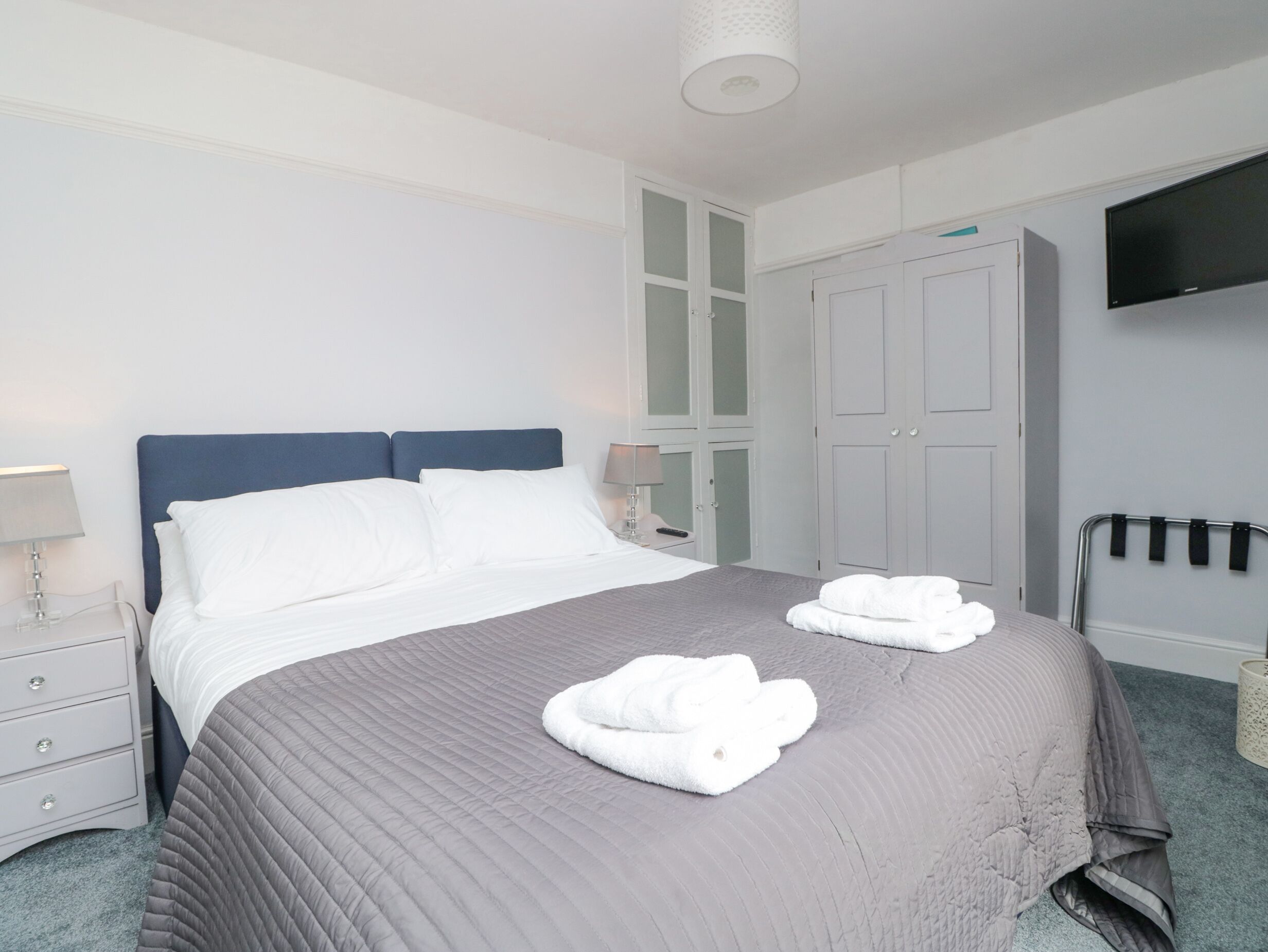 Een net opgemaakt bed met een grijze deken, witte kussens en twee gevouwen witte handdoeken. Er staat een nachtkastje met een lamp, een garderobe met glazen deuren en een flatscreen televisie aan de muur bevestigd.
