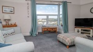 Una sala de estar con un sofá, un reposapiés y una televisión sobre un mueble. Hay una ventana con cortinas, una pequeña mesa con libros y una lámpara en una mesita.