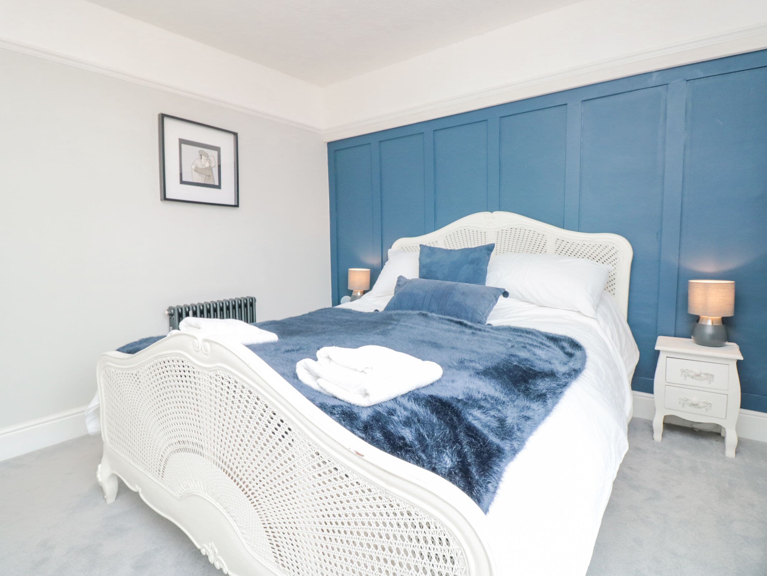Un dormitorio con cama blanca, ropa de cama azul, radiador blanco y un cuadro enmarcado en la pared.