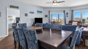 A dining area with a large table, blue upholstered chairs, and a ceiling fan. - Whispering Waves: Ocean View Condo With Covered Balcony (Oak Island)