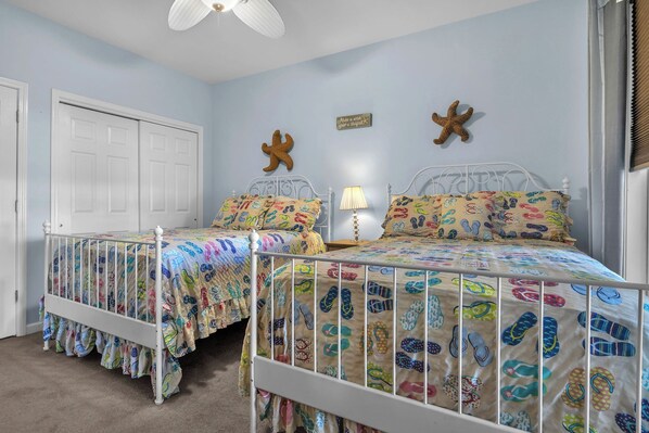 Quarto com duas camas, cada uma com um cobreleiro colorido com estampas de chinelos. Há decorações de estrelas-do-mar na parede, uma porta e um ventilador de teto.