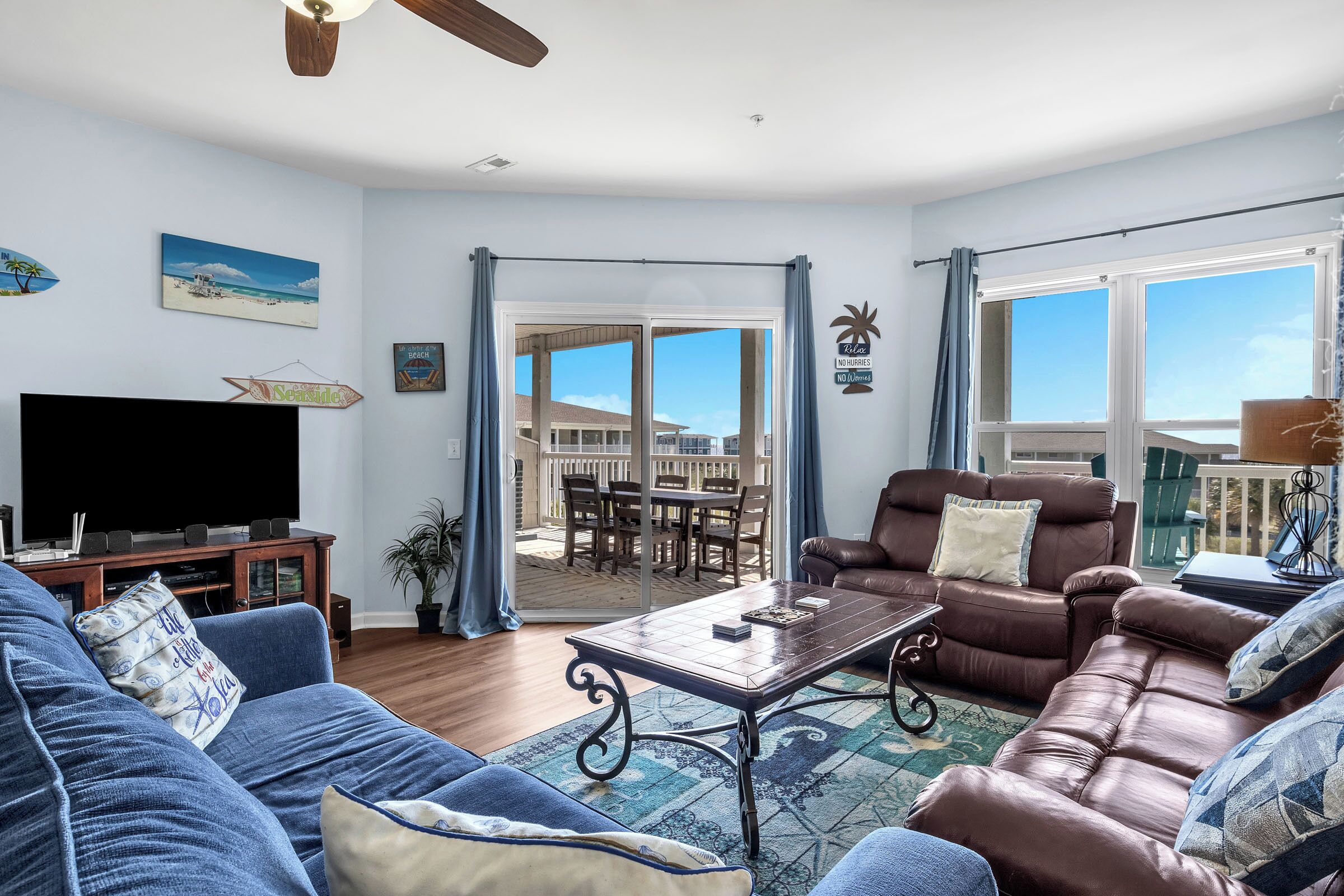 A living room with a large flat-screen TV, a brown leather sofa, a blue couch, and a coffee table. There is a sliding glass door leading to an outdoor patio area with a view of the ocean.