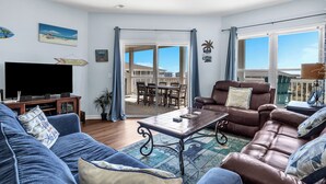 A living room with a large flat-screen TV, a brown leather sofa, a blue couch, and a coffee table. There is a sliding glass door leading to an outdoor patio area with a view of the ocean. - Whispering Waves: Ocean View Condo With Covered Balcony (Oak Island)