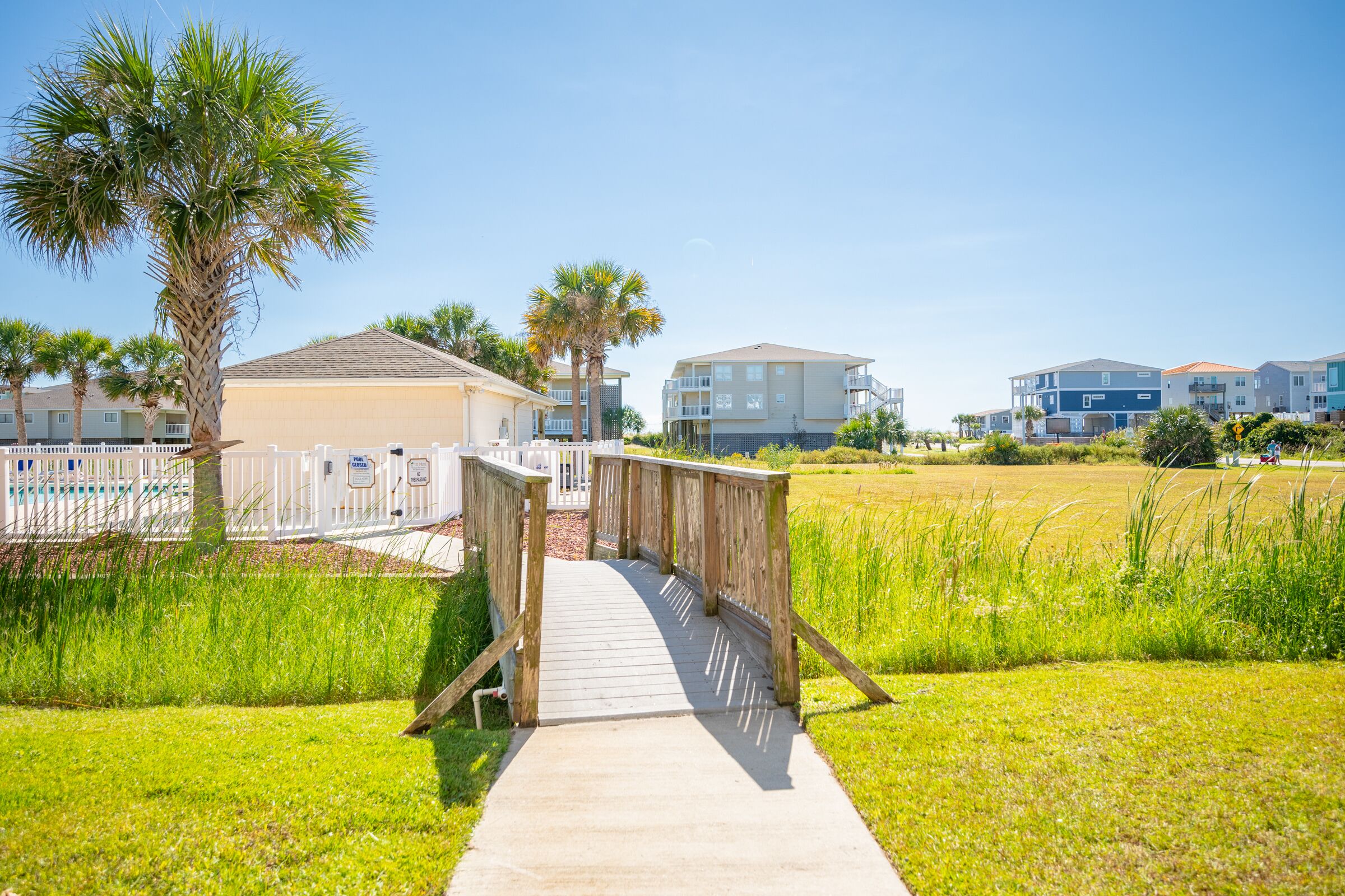 A wooden boardwalk leading to a building with a white fence and palm trees.