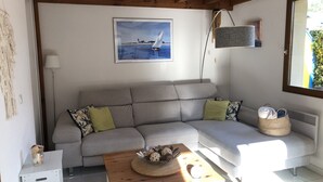 A cozy living room with a grey sofa, a small white side table, a wooden coffee table with decorative items, a framed painting of a sailboat, and a window with a view of greenery.