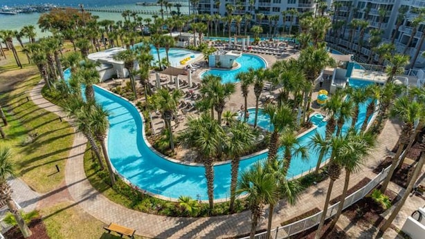 Ein Resort mit einem großen, geschwungenen Pool, umgeben von Palmen und Liegestühlen, mit Blick auf den Ozean und einem Pier im Hintergrund.