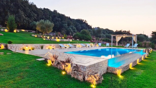 Una piscina rectangular amb un solàrium de pedra, envoltada per un jardí ben cuidat i parets de pedra.