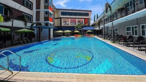 A swimming pool with a circular design, surrounded by lounge chairs and umbrellas, with buildings in the background. - Inter Park Hotel (Olongapo)