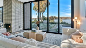 Una sala de estar moderna con chimenea, ventanas amplias y vista a un lago y árboles.