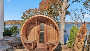Estructura de sauna de madera con puerta y dos bancos, ubicada junto a un lago rodeado de follaje otoñal.