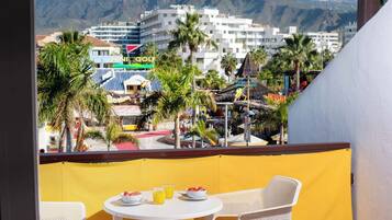 A balcony with a yellow partition, white wicker furniture, and a table with breakfast items. In the background, there are buildings, palm trees, and a mountainous landscape.