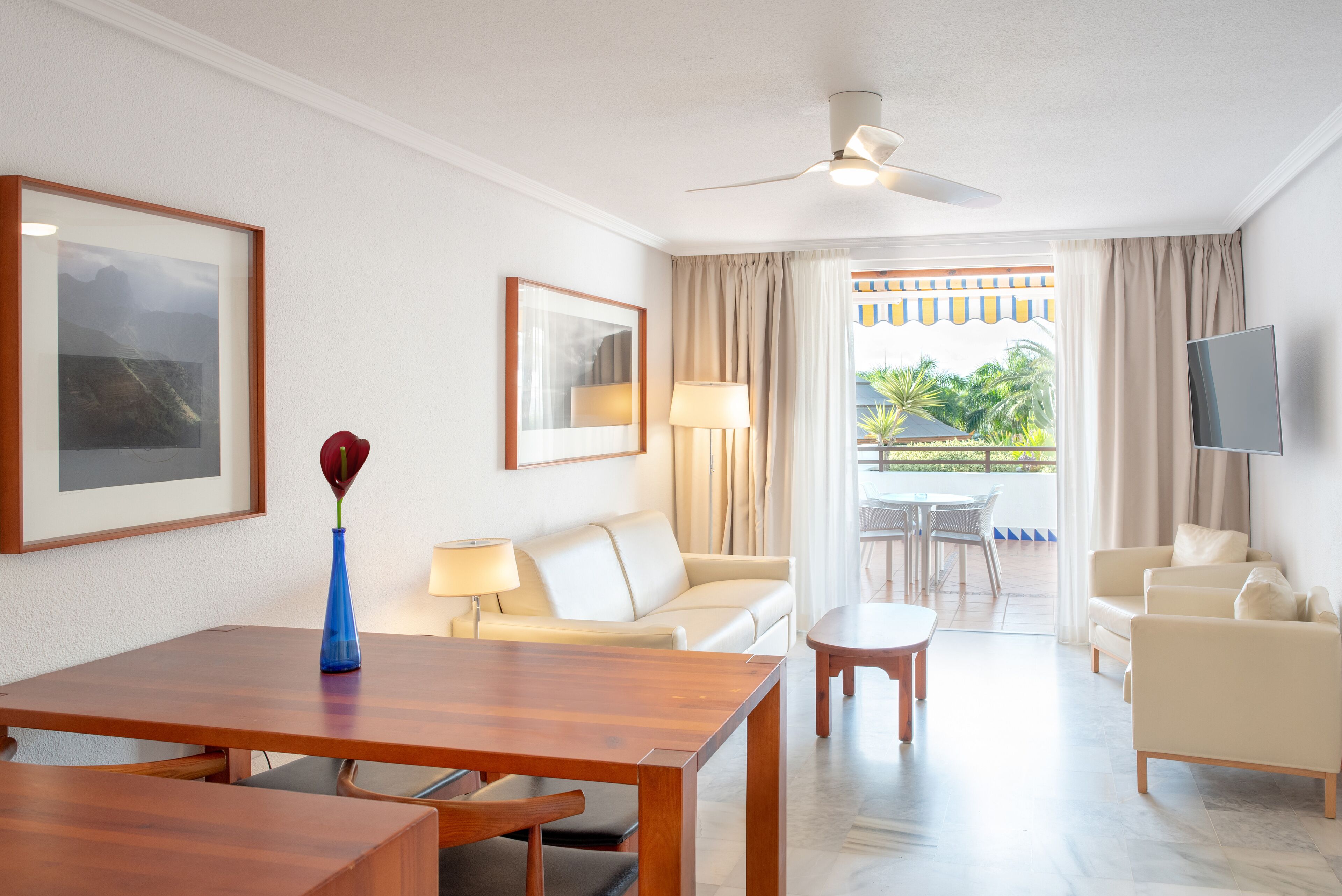 Habitación de hotel con una mesa de comedor de madera, un sofá, un televisor y un balcón con mobiliario exterior.