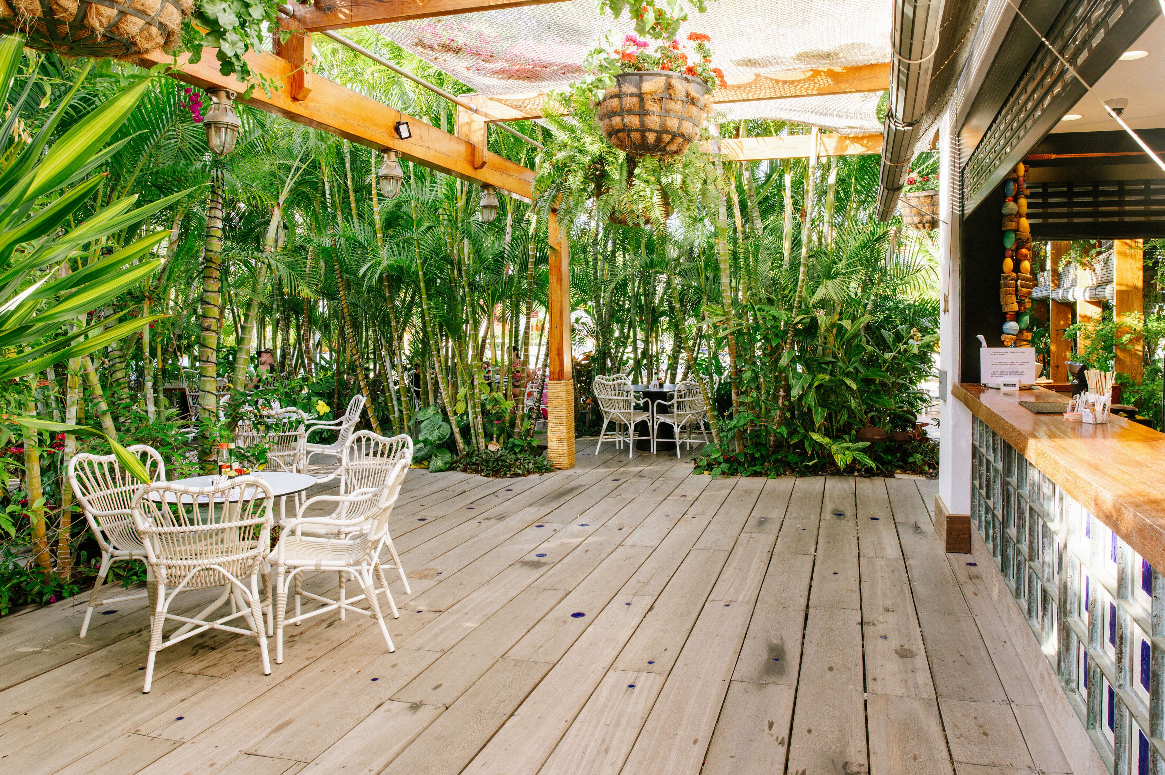 Un patio in legno con mobili in vimini bianco, immerso in piante tropicali rigogliose e un vaso sospeso fiorito.