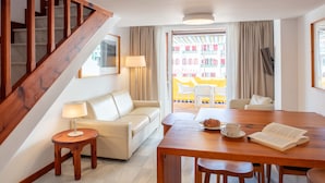 A hotel room with a wooden staircase, a sofa, a dining table with chairs, a lamp, and a view of a colorful outdoor area. - Aparthotel Parque Santiago III (Arona)