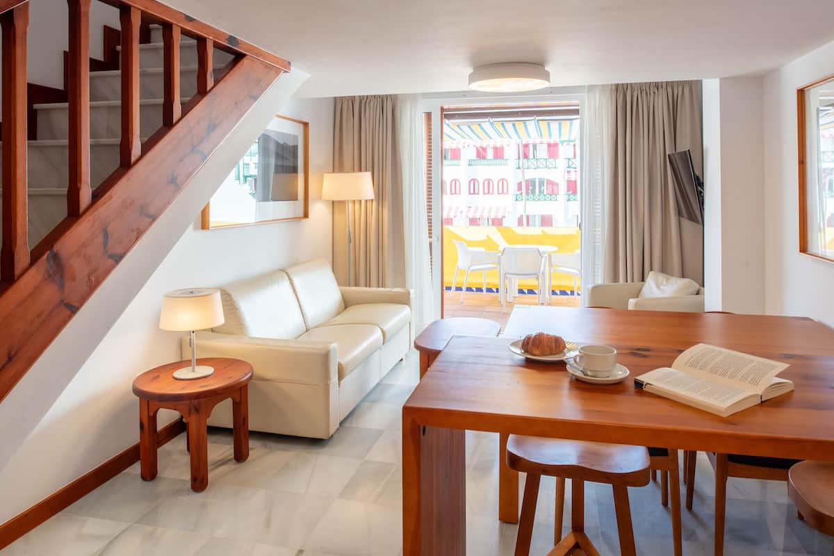 Quarto de hotel com escada de madeira, um sofá, mesa de jantar com cadeiras, uma luminária e vista para uma área externa colorida.