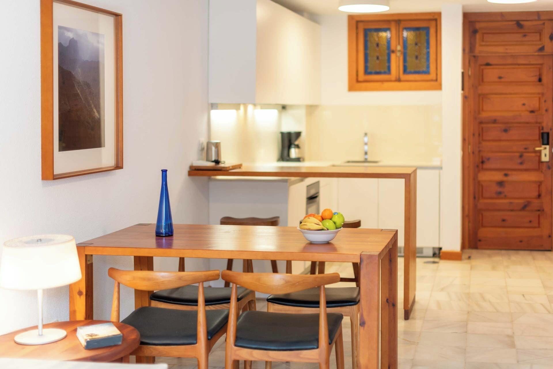 A dining area with a wooden table and chairs, a bowl of fruit, and a framed picture on the wall.