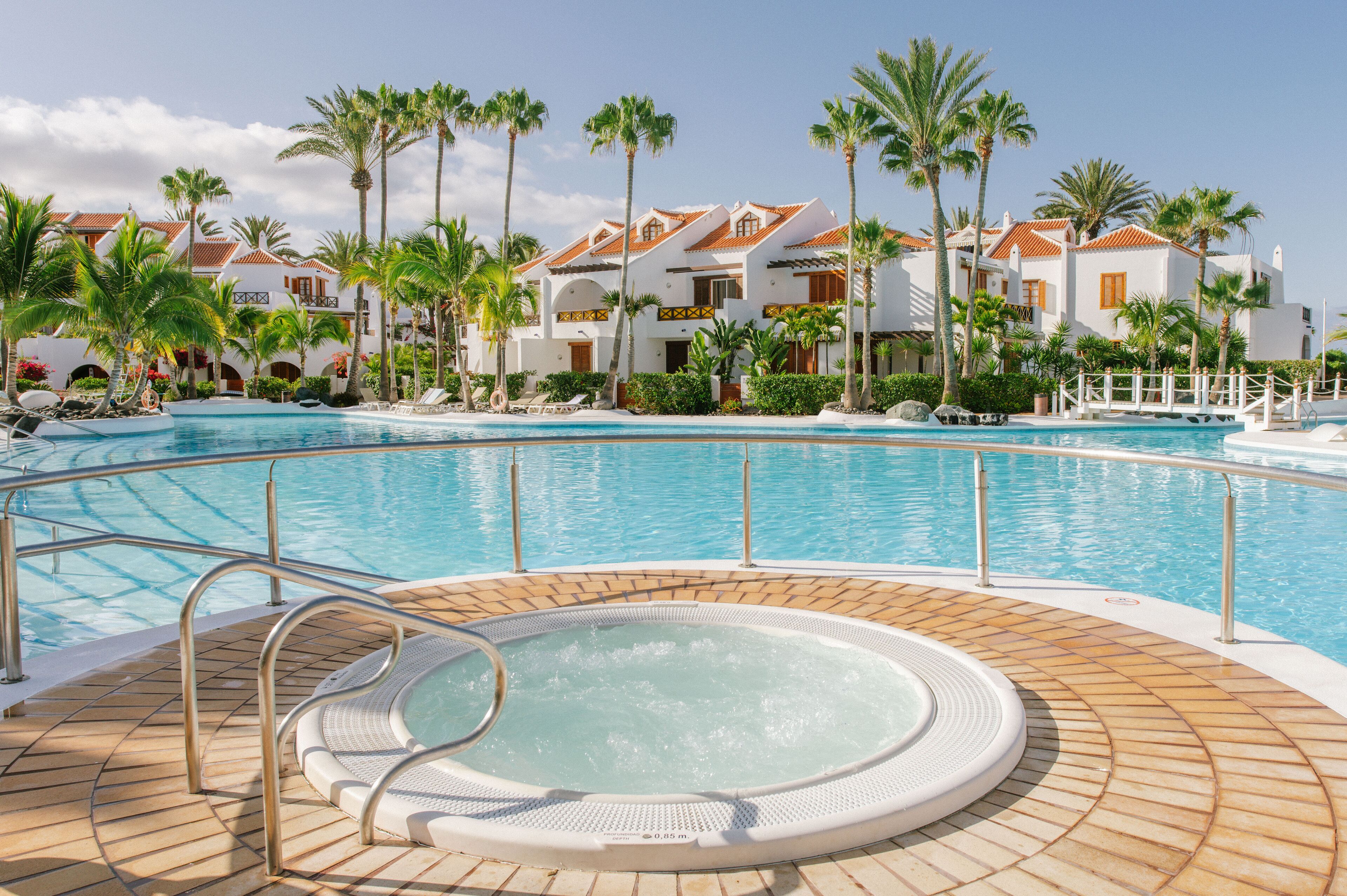 Zona de piscina en un resort con jacuzzi, palmeras y un edificio con techo de tejas.