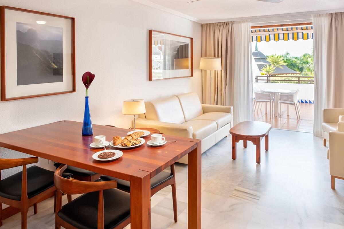Quarto de hotel com mesa de jantar de madeira, um sofá, uma mesinha redonda e varanda com vista para vegetação e um edifício.