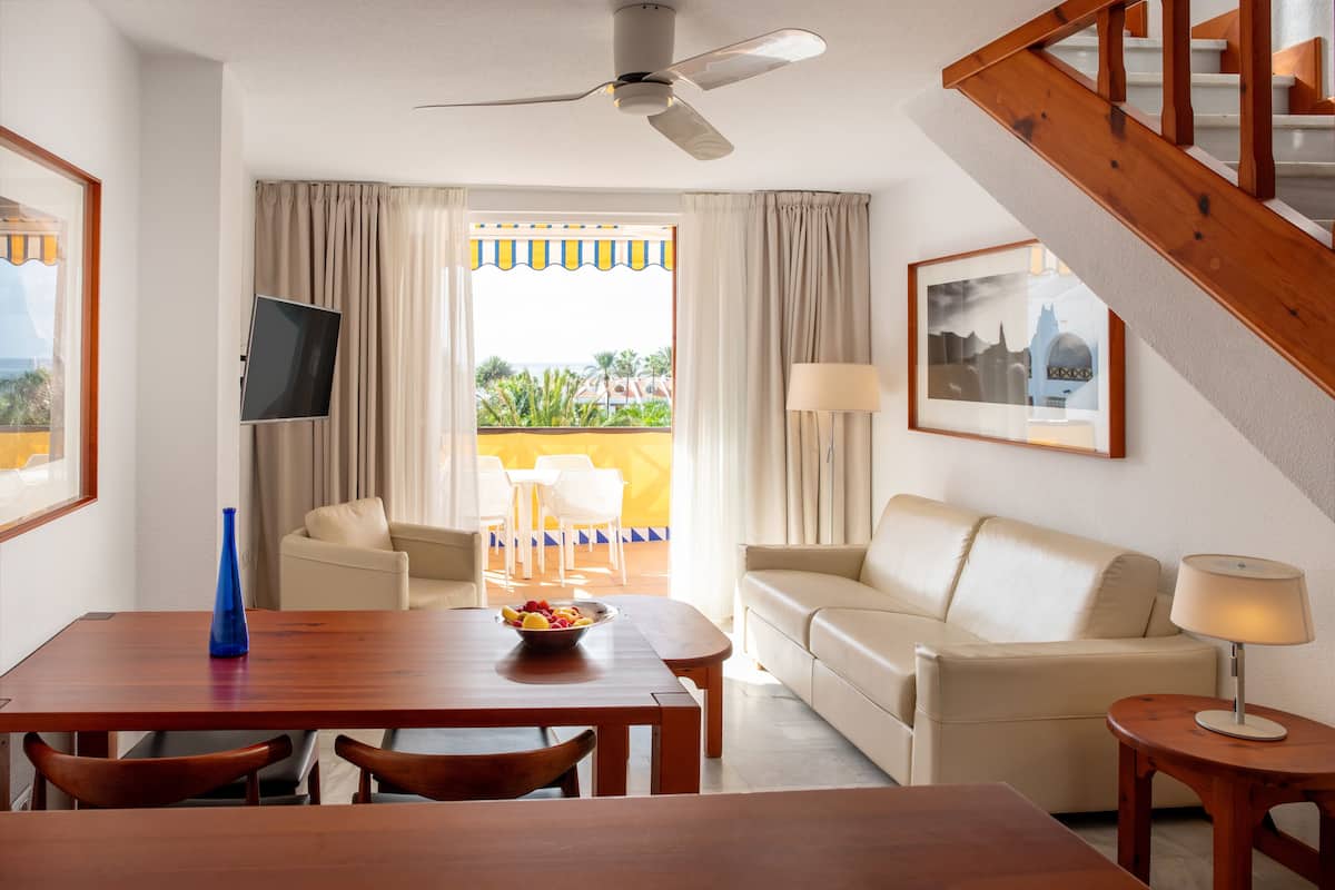 Quarto de hotel com mesa de jantar de madeira, sofá bege e televisão na parede.