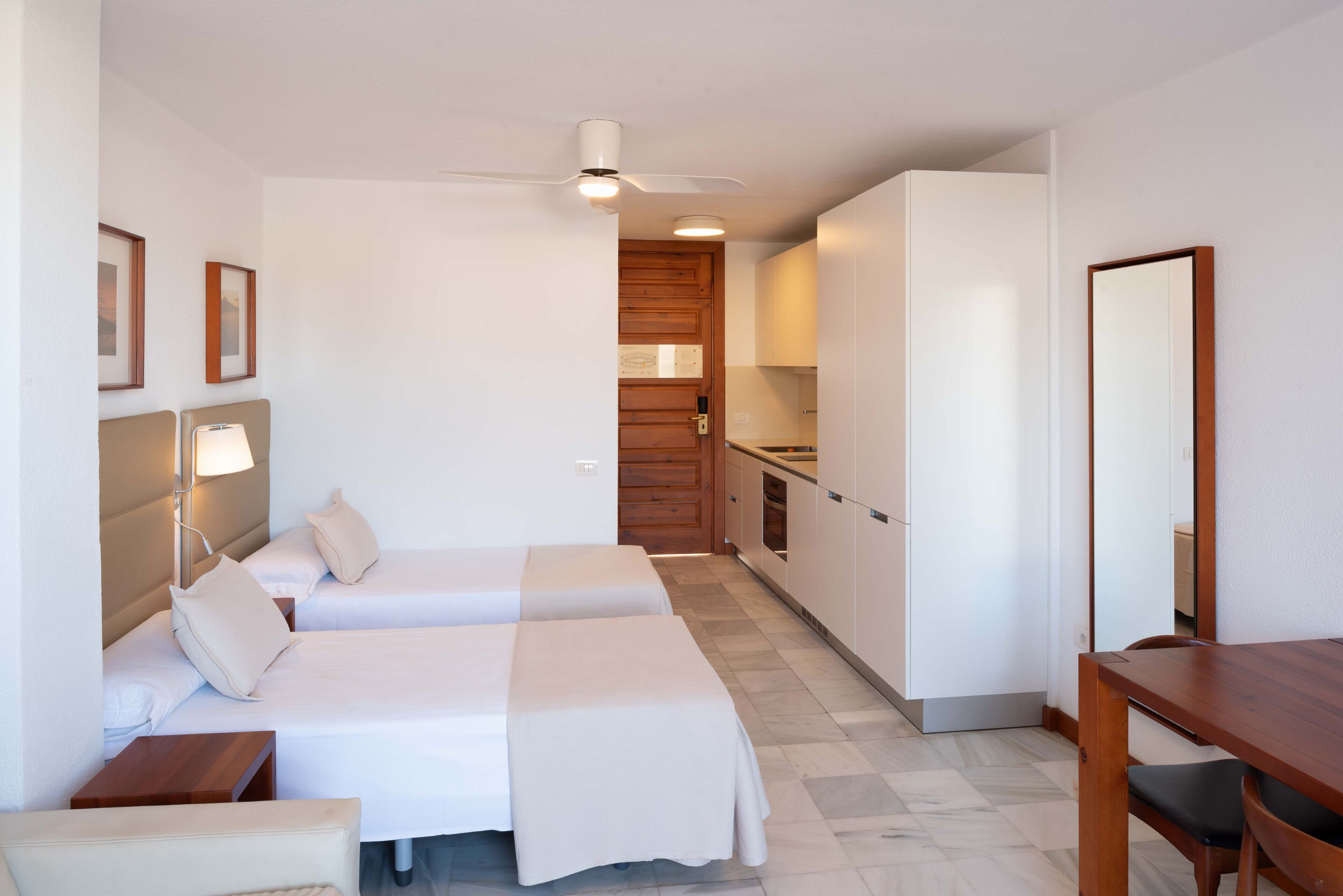 A hotel room with two beds, a desk, a chair, a ceiling fan, and a kitchenette.