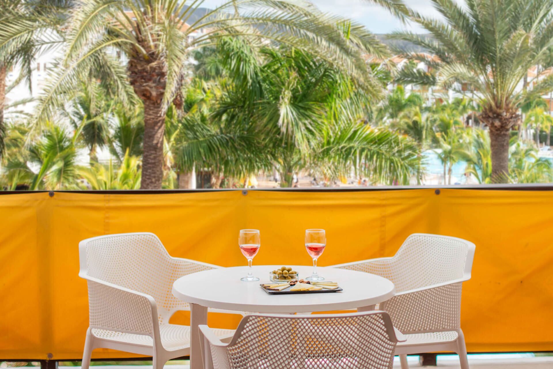 A round table with two white chairs, two glasses of red wine, and a small bowl of olives.