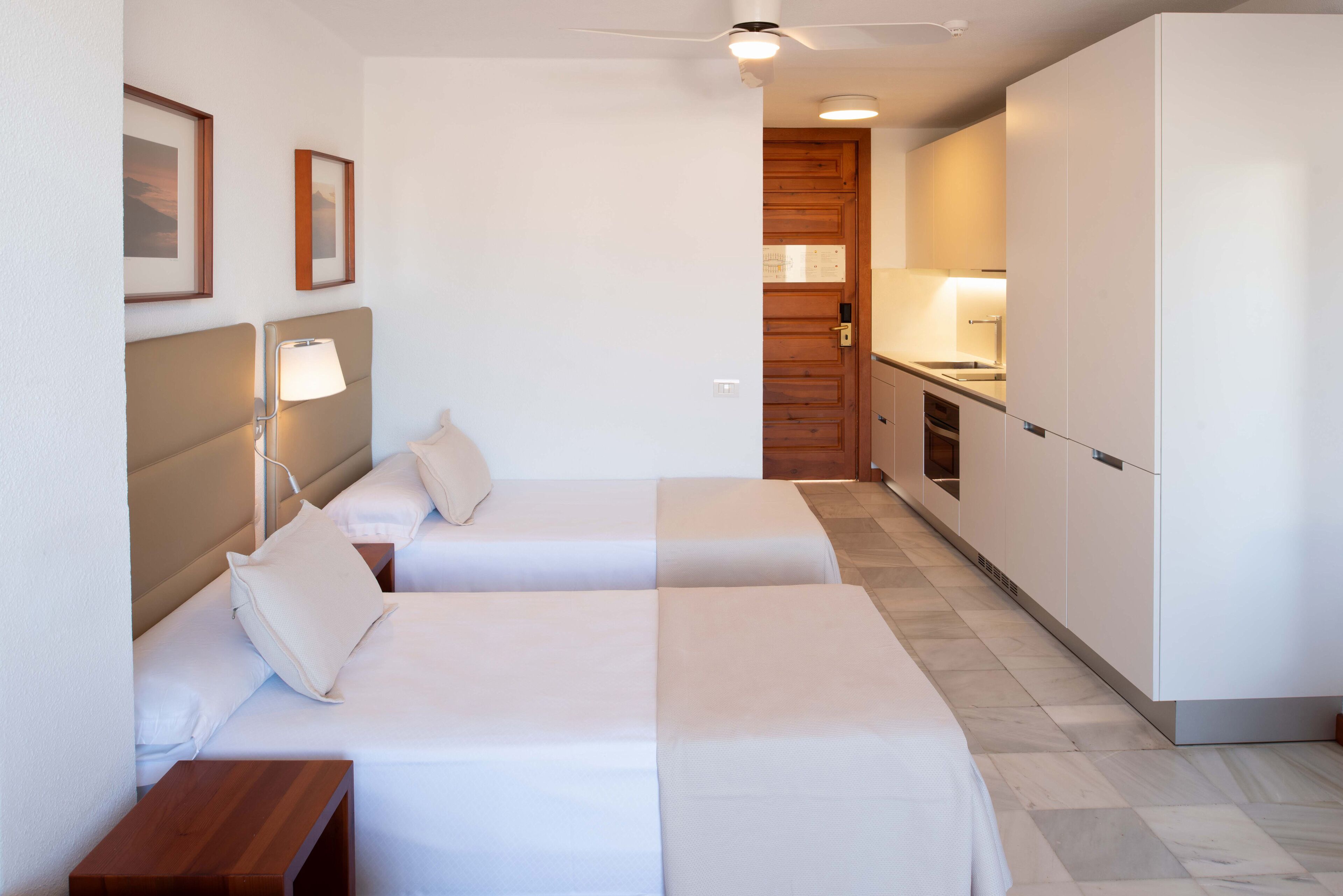 A compact hotel room with two beds, a kitchenette, and a ceiling fan.