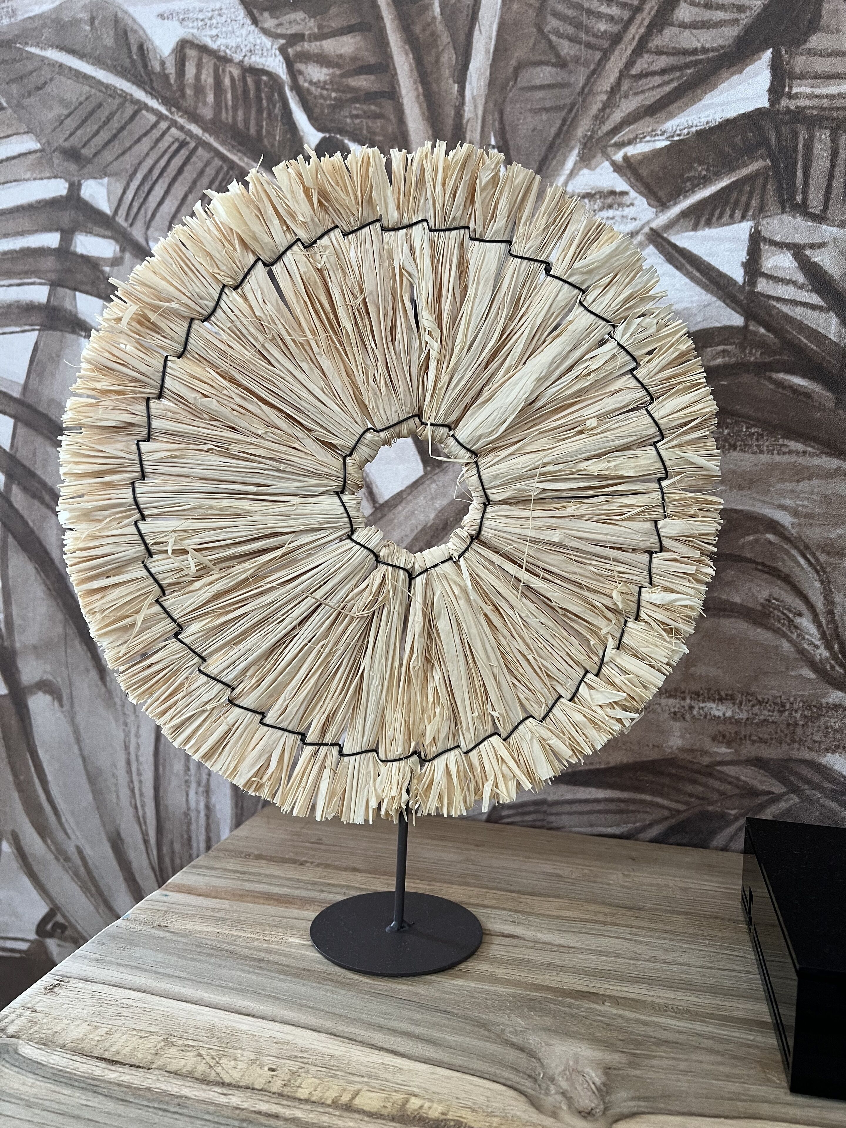 Un objeto circular tejido, similar al de paja, con una superficie reflectante en el centro, montado sobre un soporte negro, colocado sobre una superficie de madera con fondo decorado.