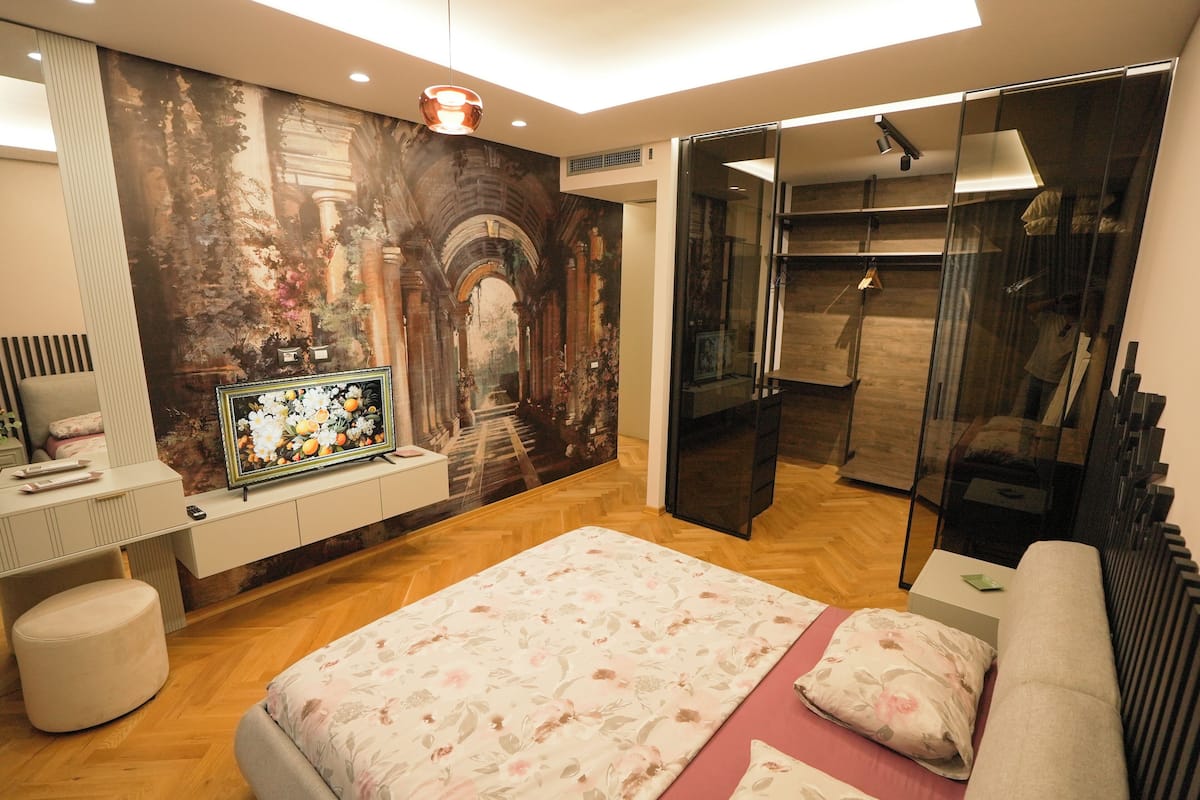 A modern bedroom with a large bed, a TV, a wardrobe, and a glass door leading to a walk-in closet.