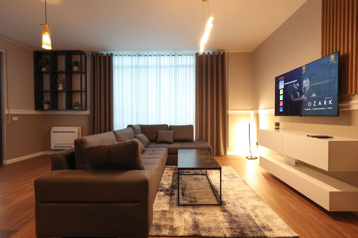 A modern living room with a sectional sofa, a flat-screen TV, a built-in shelf, and a large window with curtains.