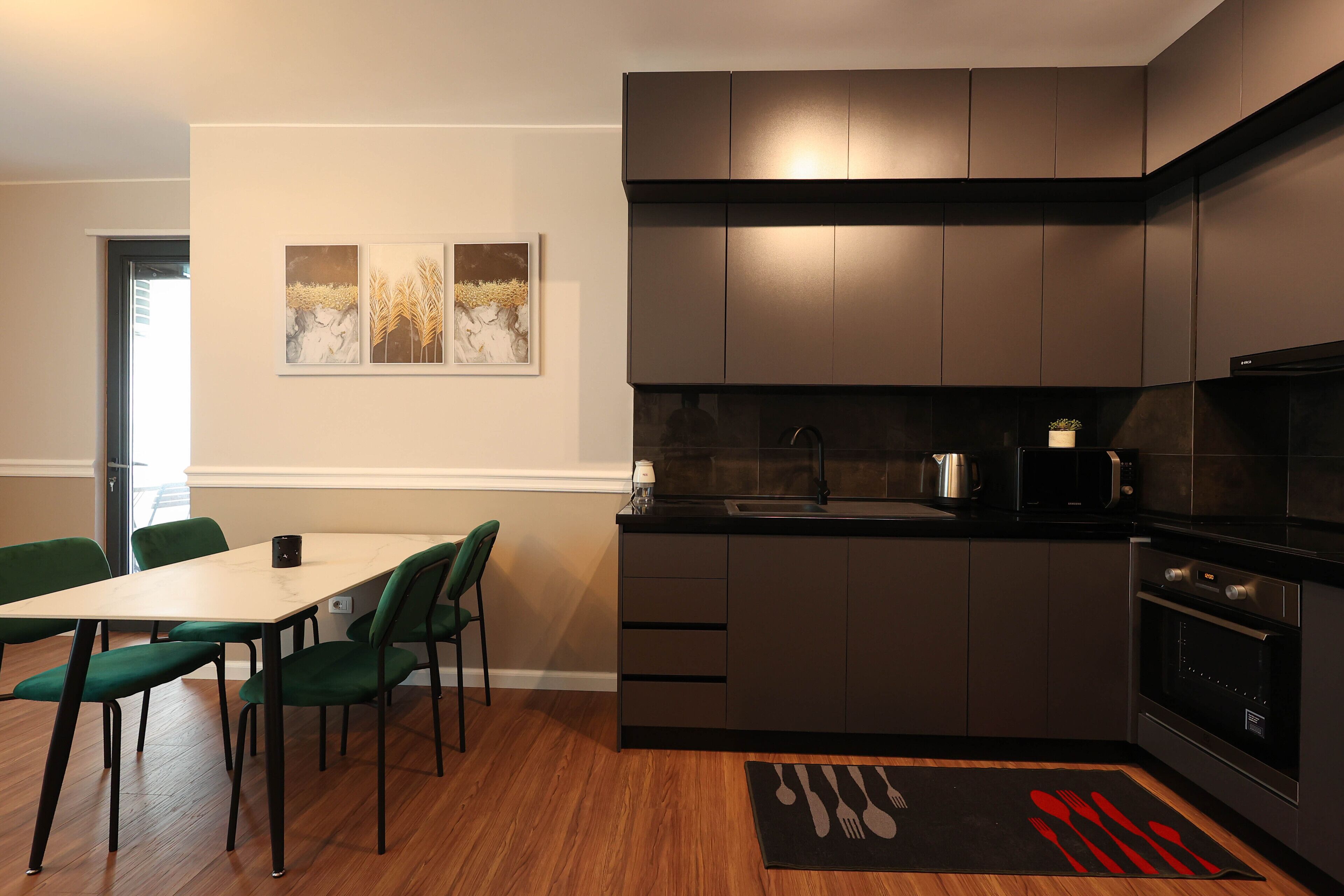A modern kitchen with dark cabinetry, a wooden dining table with green chairs, and a black rug with cutlery design.