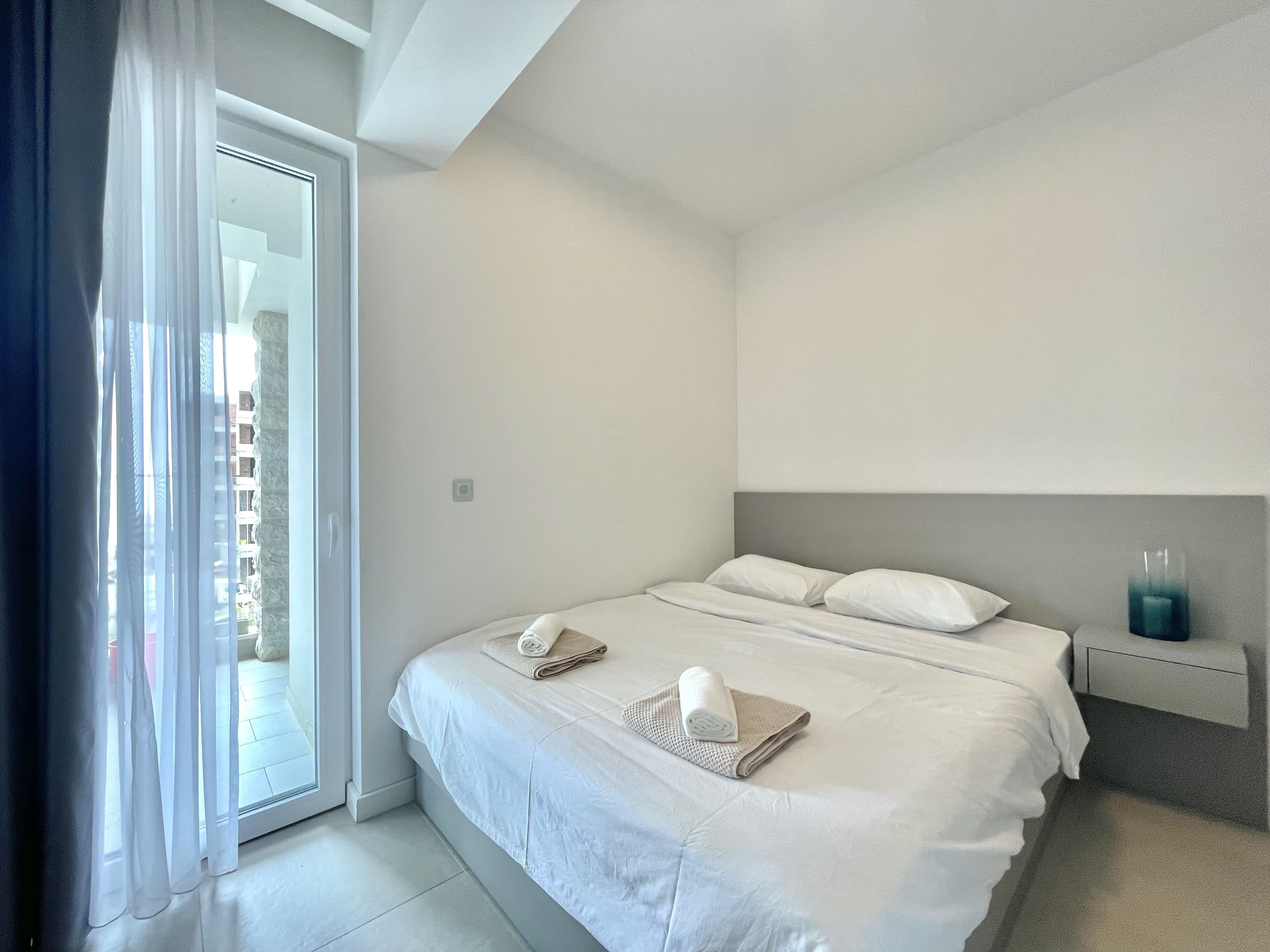 A modern hotel room with a large bed, a nightstand, and a view through a glass door.