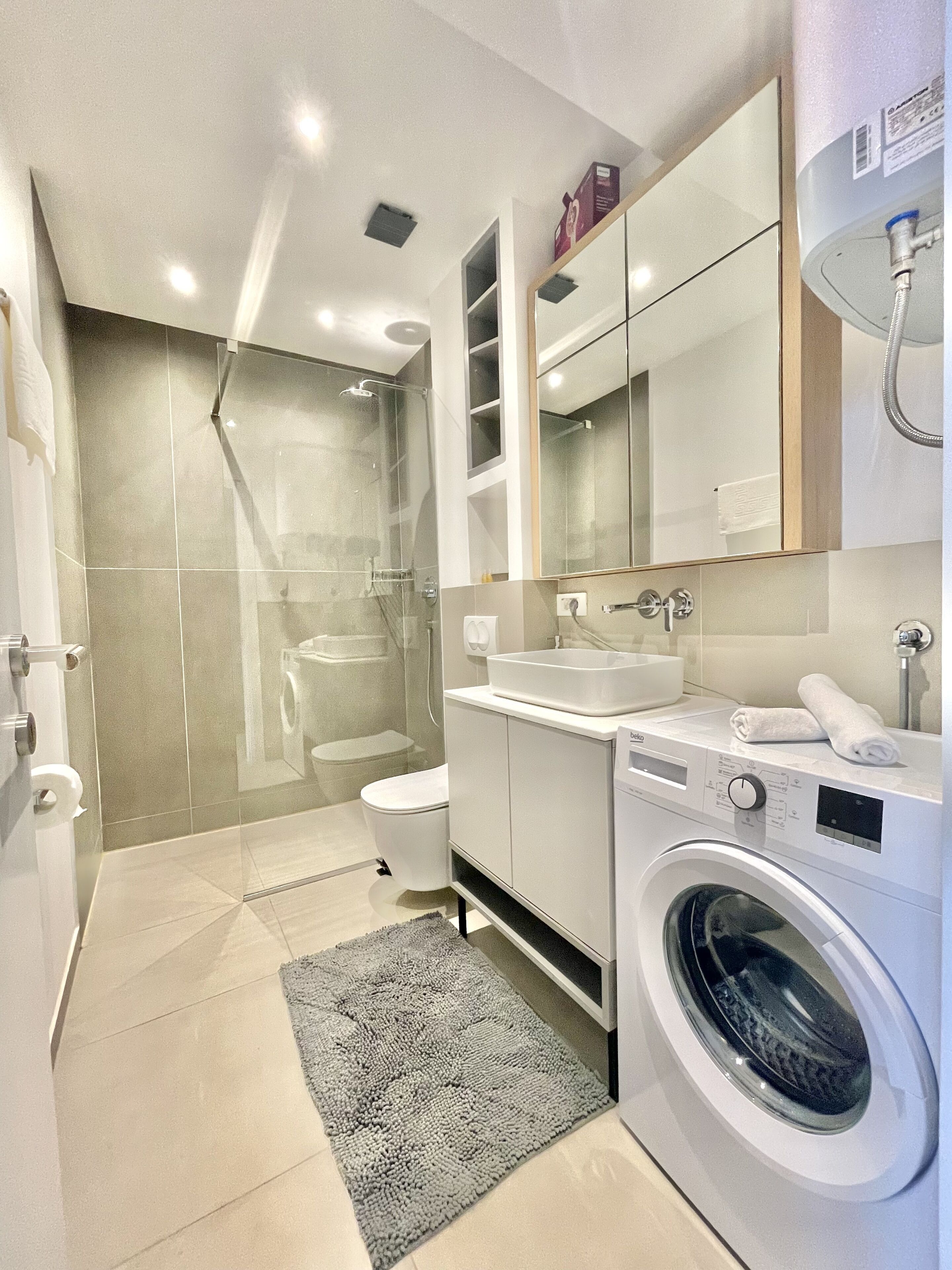 A modern bathroom with a glass-enclosed shower, a toilet, a sink, a mirror, and a washing machine.