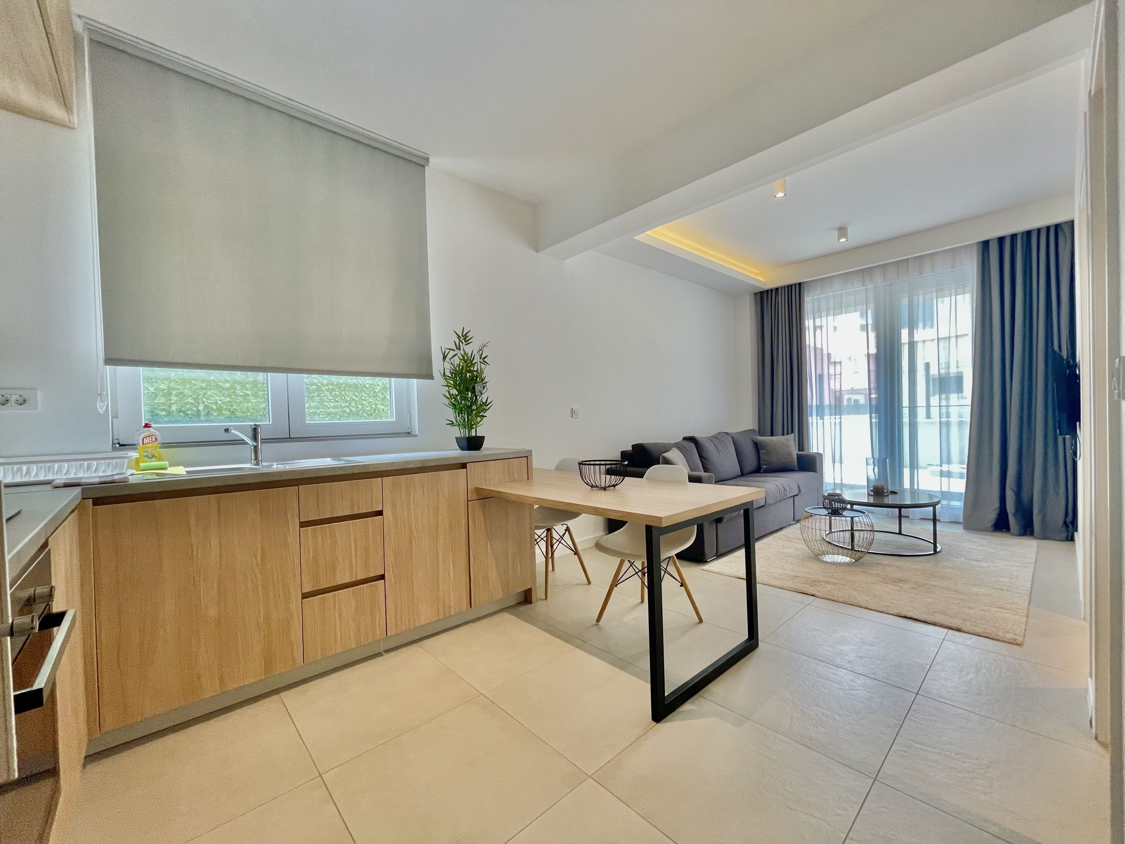 A modern kitchen with wooden cabinets, a dining table, and a sofa in a living area with large windows and a view of the outdoors.