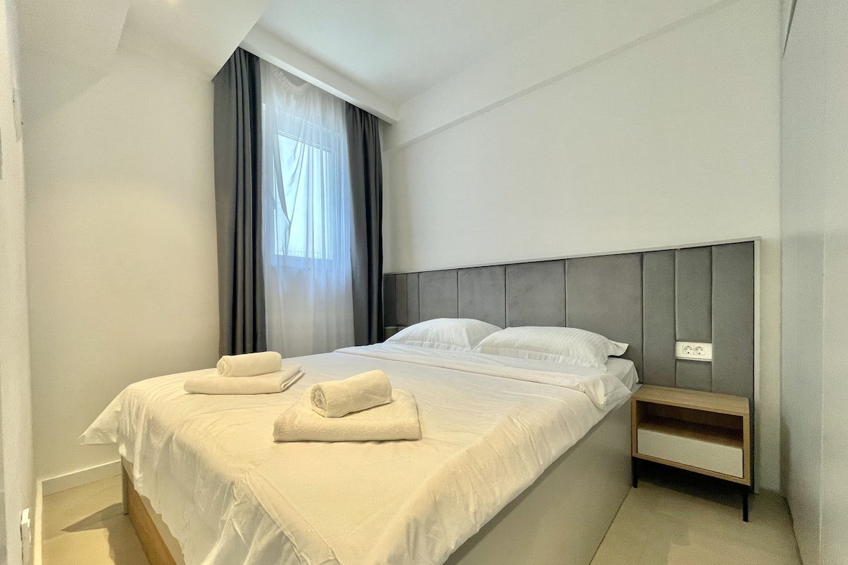 A neatly made bed with white linens and two rolled towels, a bedside table, and a window with sheer curtains.