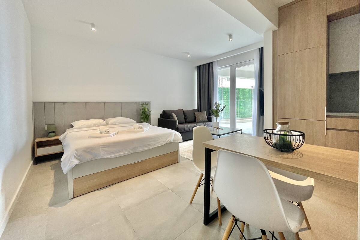 A modern hotel room with a bed, a sofa, a dining table, and a kitchen area.