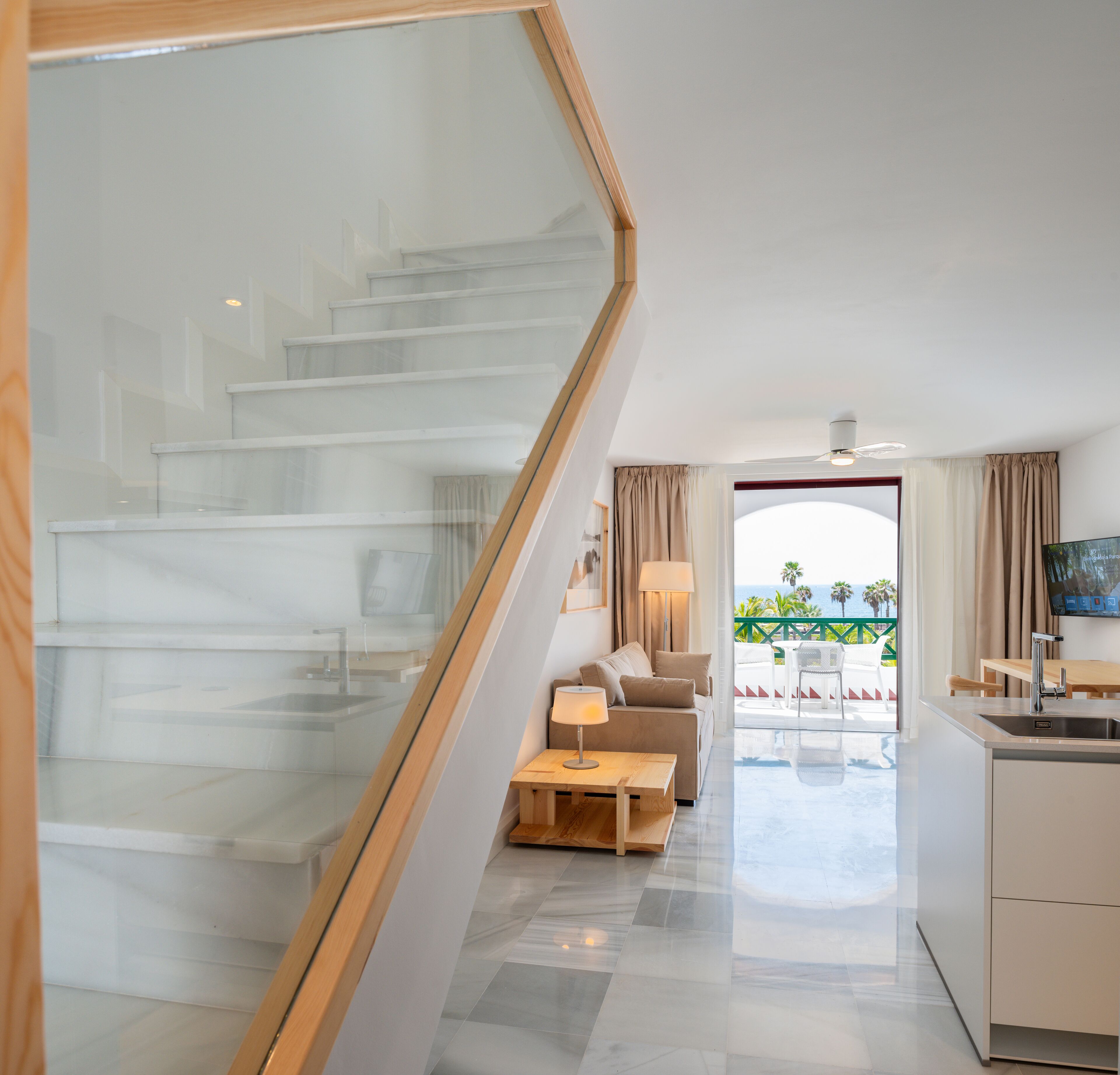 A modern hotel room with a glass staircase, a living area with a sofa and coffee table, a kitchen with a sink, and a view of the ocean.