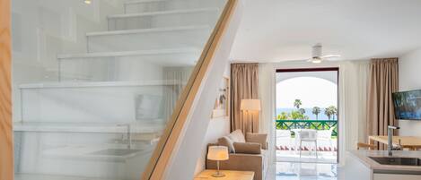 A modern hotel room with a glass staircase, a living area with a sofa and coffee table, a kitchen with a sink, and a view of the ocean.