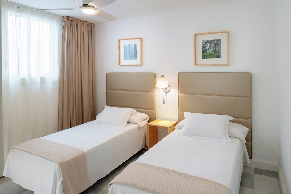 A hotel room with two single beds, a ceiling fan, and two framed pictures on the wall.