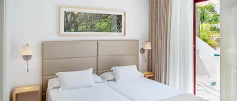 A double bed with white linens and a beige headboard, two bedside tables with lamps, a framed picture on the wall, and a view of greenery outside.