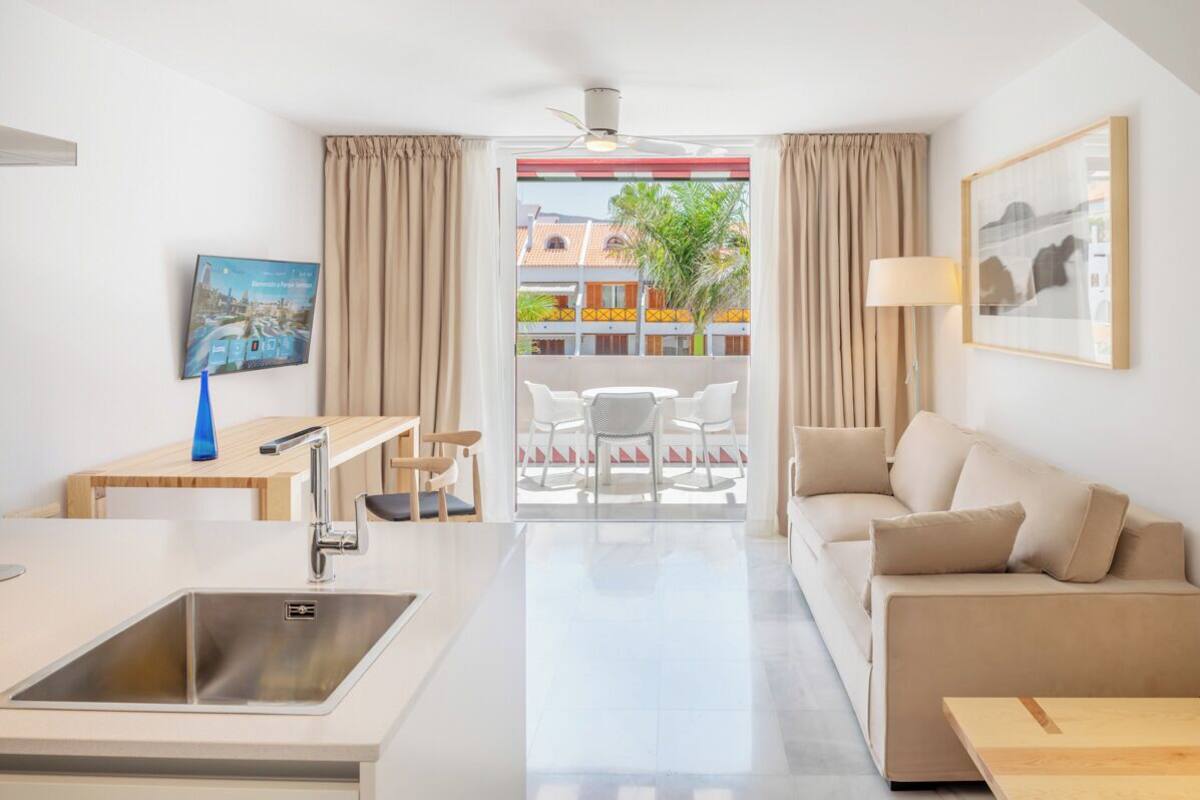 A modern hotel room with a kitchenette, a dining area, a sofa, and a balcony with a view of buildings and a palm tree.