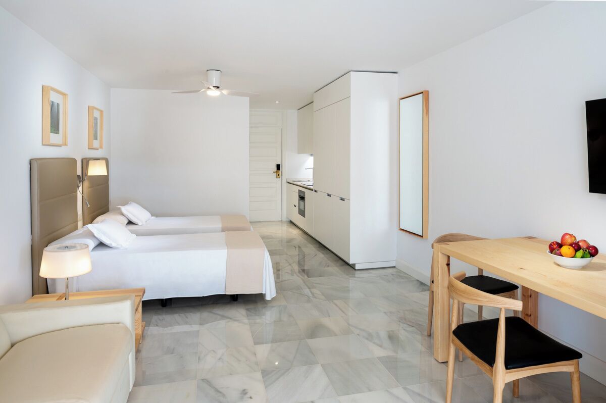 A modern hotel room with a bed, bedside table, lamp, and a dining area with a table and chairs.