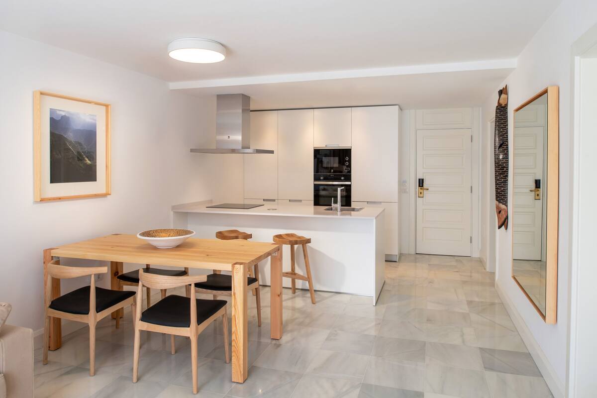 A modern kitchen with a central island, white cabinetry, and a built-in oven. There is a dining area with a wooden table and chairs, and a framed picture on the wall.