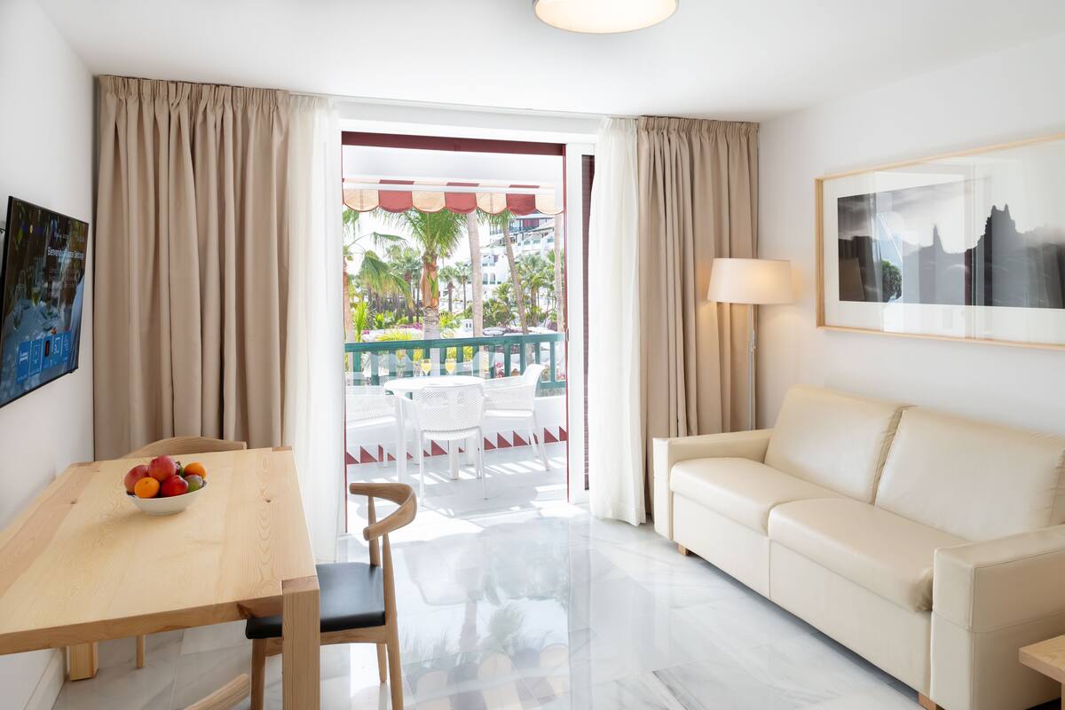 A hotel room with a wooden dining table, a bowl of fruit, a white sofa, and a balcony with outdoor seating.
