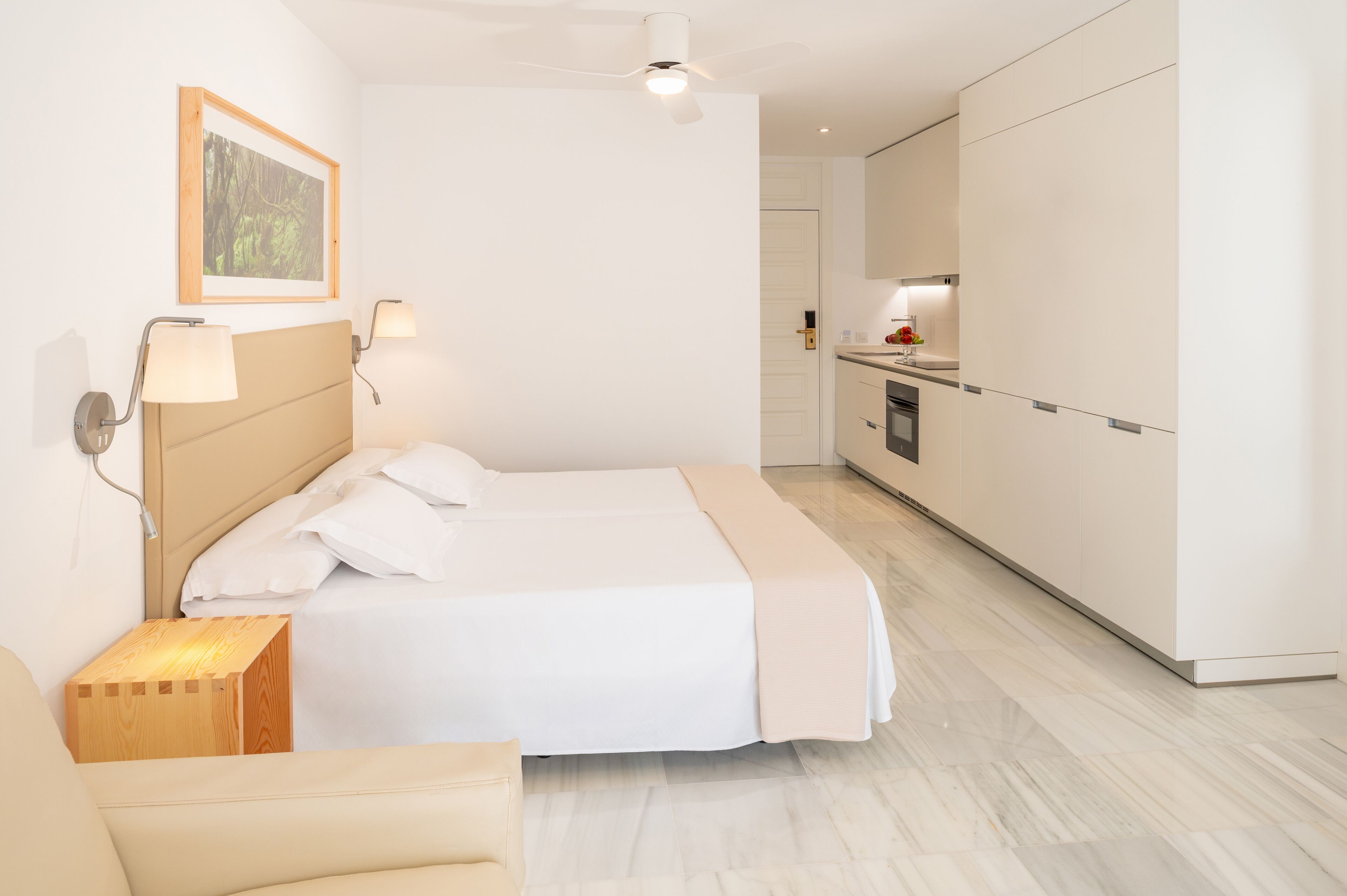 Ein modernes Hotelzimmer mit einem großen Bett, Nachttischlampen, einem Holznachttisch, einem kleinen Holzschemel und Blick in den angrenzenden Küchenbereich.