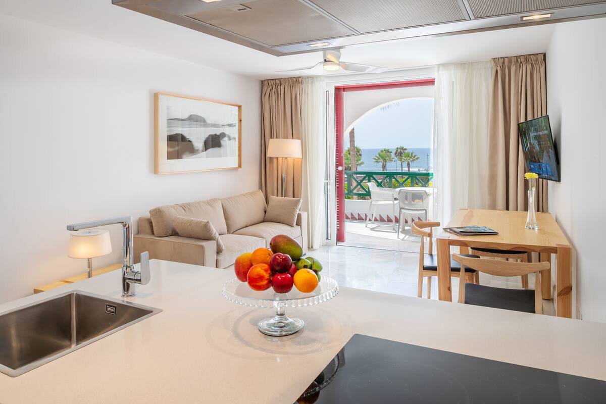 A modern hotel room with a kitchen area, a dining table, and a balcony with a view of the sea.