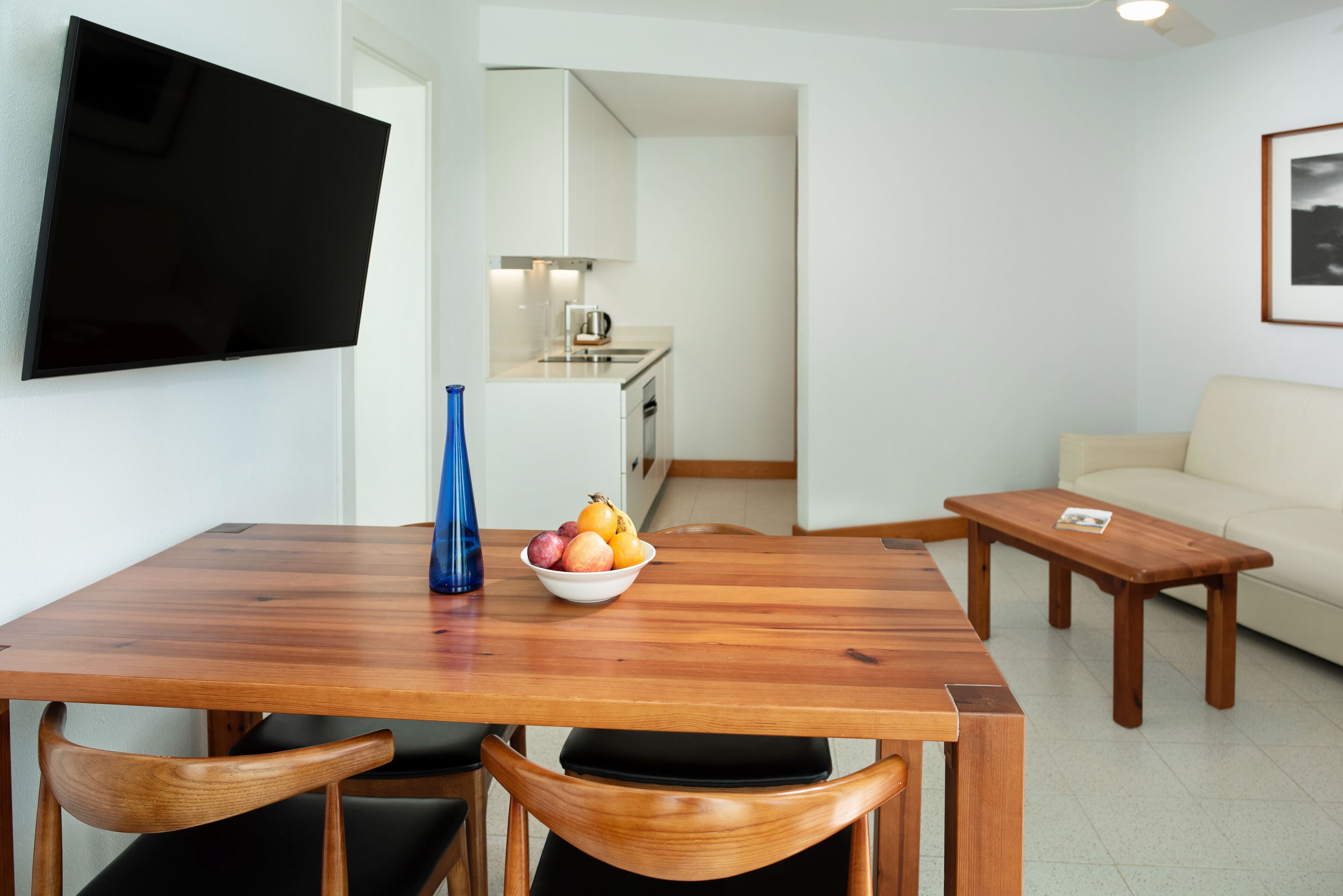 Un salon moderne avec une table à manger en bois, un téléviseur fixé au mur, un coin cuisine à l’arrière-plan et un canapé beige.